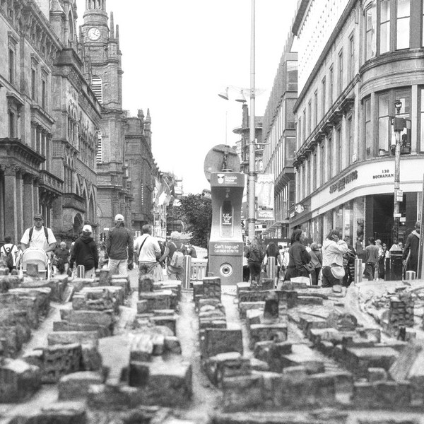 Black-and-white street scene in Glasgow city centre, with a table of densely arranged miniature building models in the foreground and pedestrians walking between tall historic commercial buildings behind.