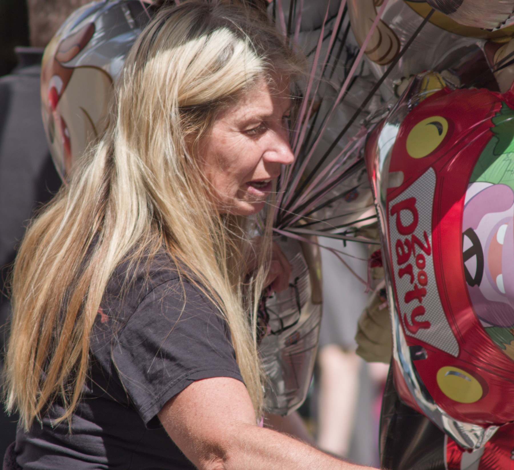Close-up of a blonde woman beside a cluster of shiny helium balloons, including a red cartoon-themed balloon.