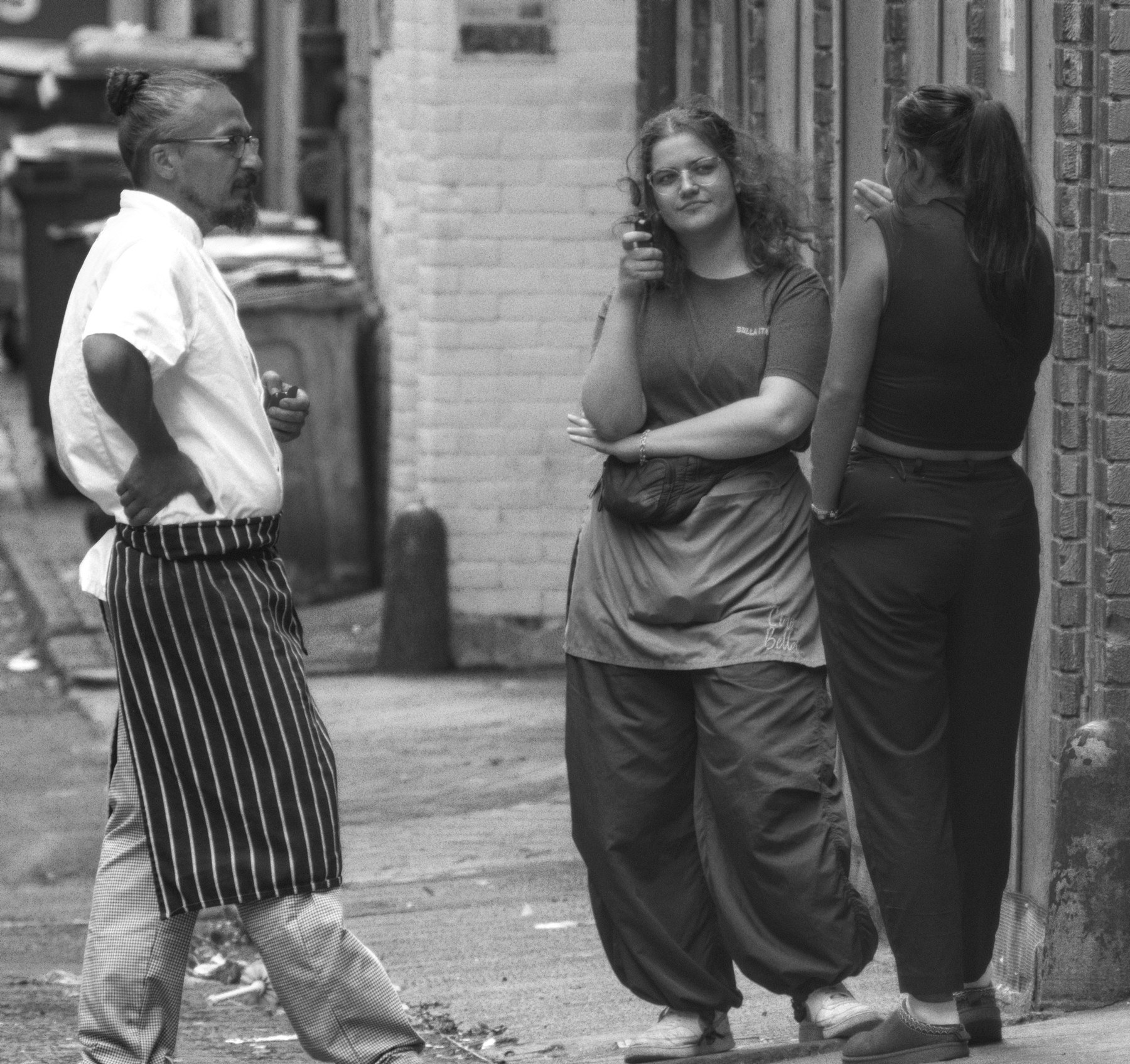 Black-and-white street scene of three people standing in a narrow city-centre lane, with one person facing the camera between two brick walls.
