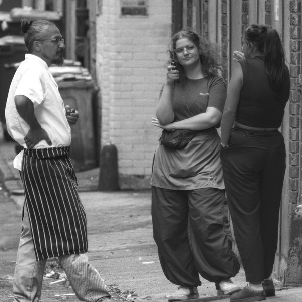 Black-and-white street scene of three people standing in a narrow city-centre lane, with one person facing the camera between two brick walls.