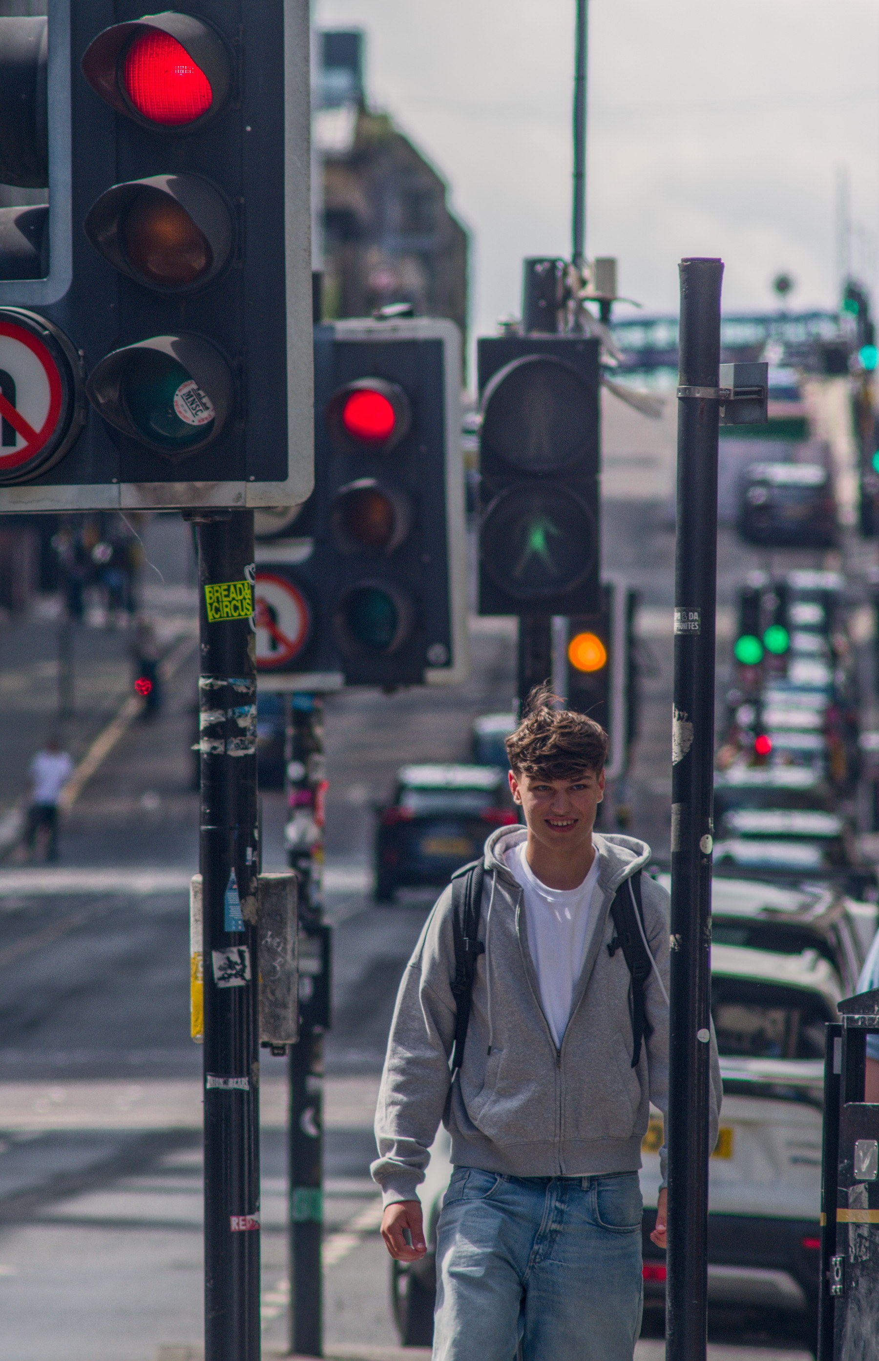 A pedestrian in a grey hoodie and backpack walks beneath red traffic lights on a busy Glasgow city-centre street, with cars and more signals receding behind him.