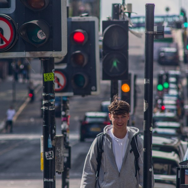 A pedestrian in a grey hoodie and backpack walks beneath red traffic lights on a busy Glasgow city-centre street, with cars and more signals receding behind him.