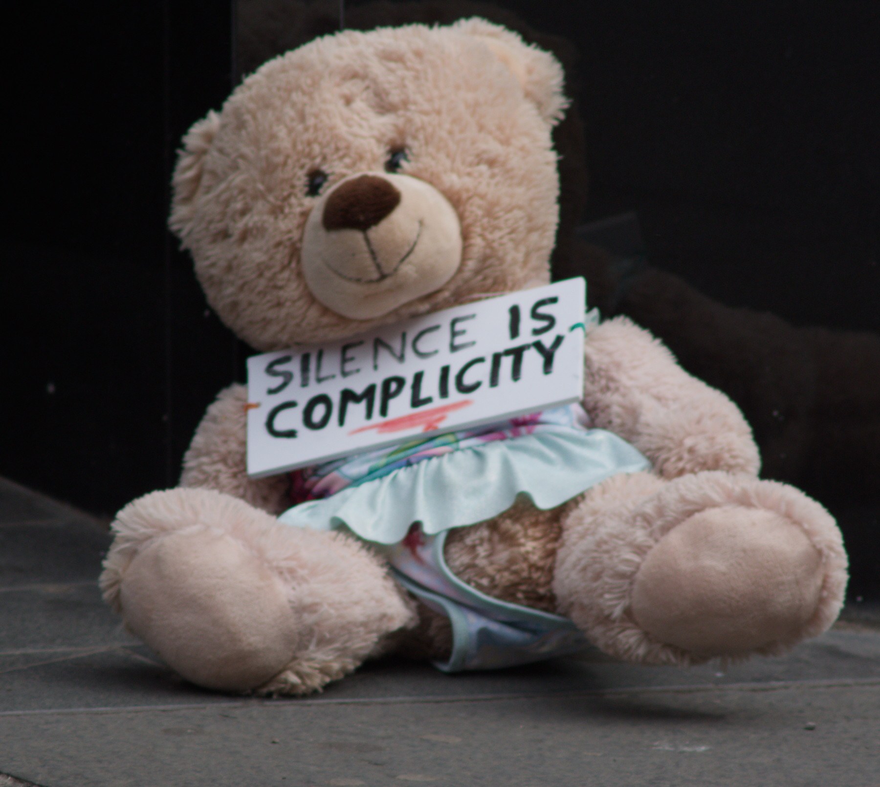 A large beige teddy bear sits on a pavement holding a handwritten sign reading “Silence is complicity.”