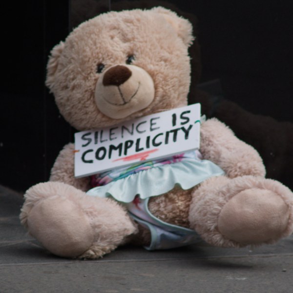 A large beige teddy bear sits on a pavement holding a handwritten sign reading “Silence is complicity.”