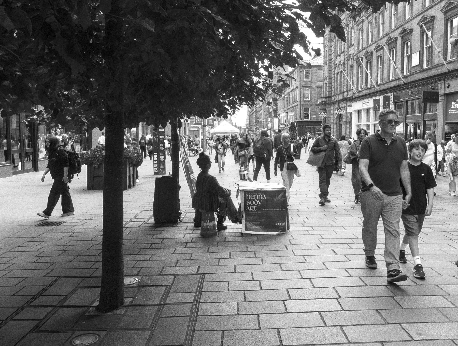 Black-and-white street scene in Glasgow city centre showing pedestrians on a broad paved shopping street, with a man and a boy walking toward the camera beneath a leafy tree.