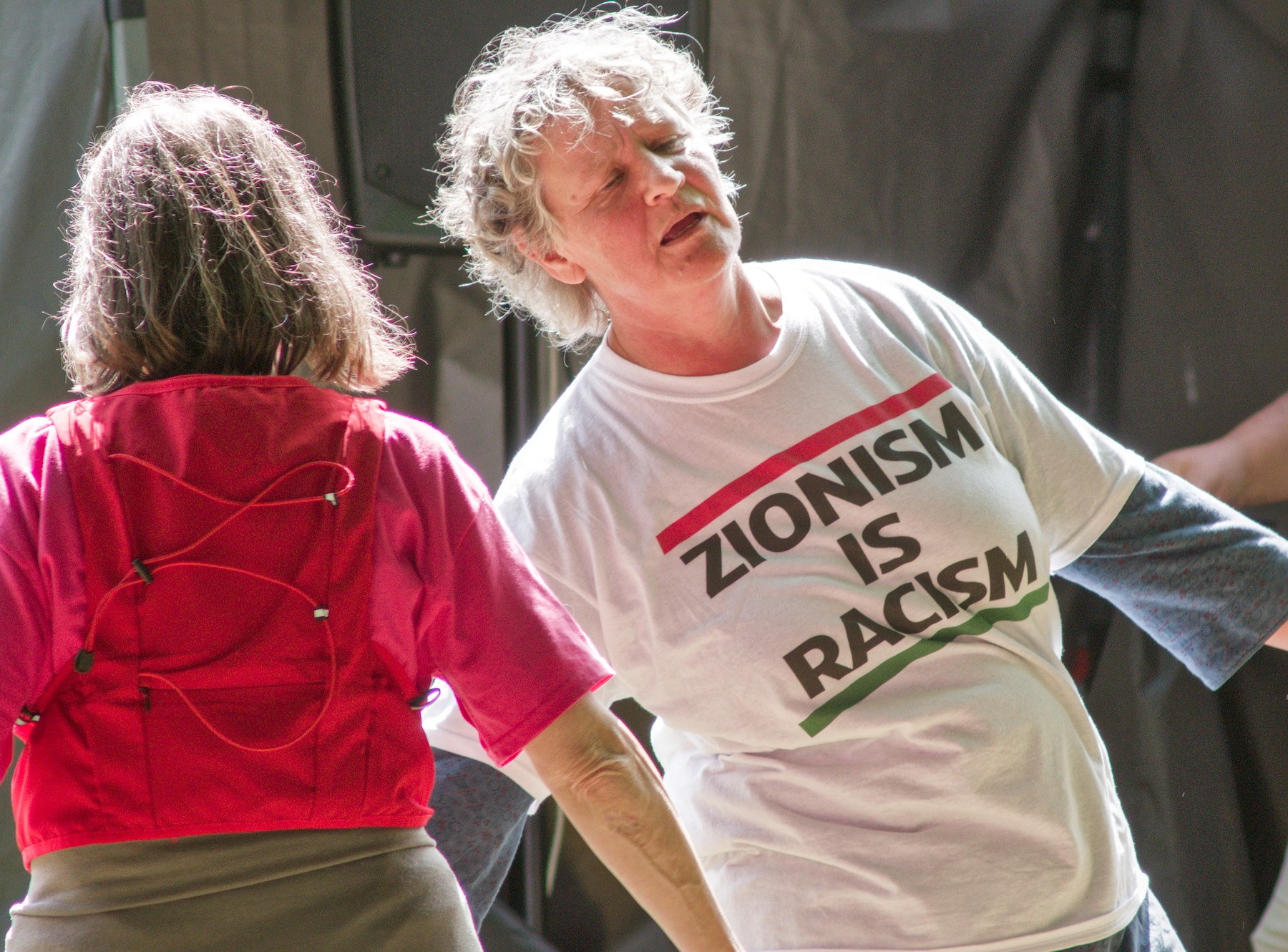Close view of a grey-haired man in a white T-shirt reading “Zionism Is Racism” beside a person seen from behind in a red top, with dark stage equipment behind them.