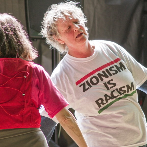 Close view of a grey-haired man in a white T-shirt reading “Zionism Is Racism” beside a person seen from behind in a red top, with dark stage equipment behind them.