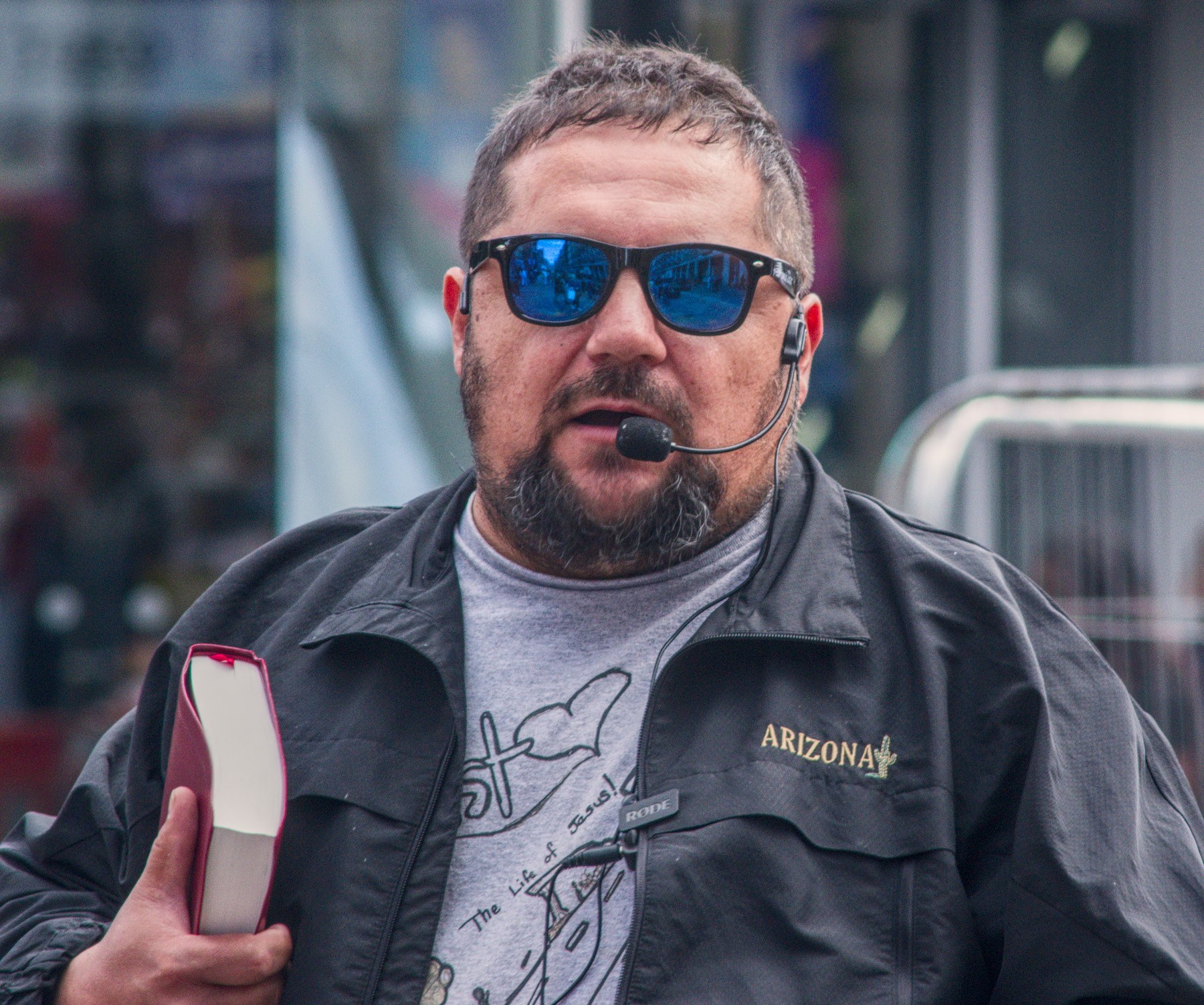 Close portrait of a man in blue mirrored sunglasses and a headset microphone, holding a red-edged book in Glasgow city centre.