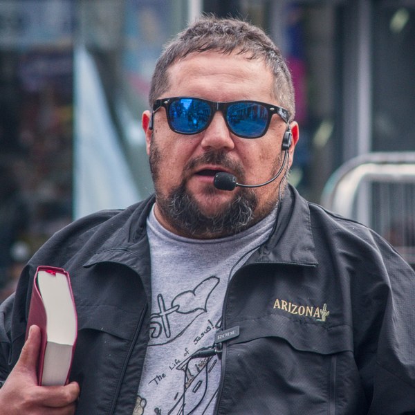 Close portrait of a man in blue mirrored sunglasses and a headset microphone, holding a red-edged book in Glasgow city centre.