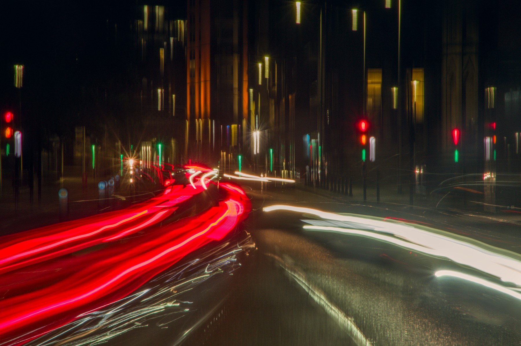 Long-exposure view down a Glasgow city street at night, with red tail-light trails curving across the left side and broad white headlight trails on the right. Red and green traffic signals glow beside dark tall buildings, and the road surface reflects the lights.
