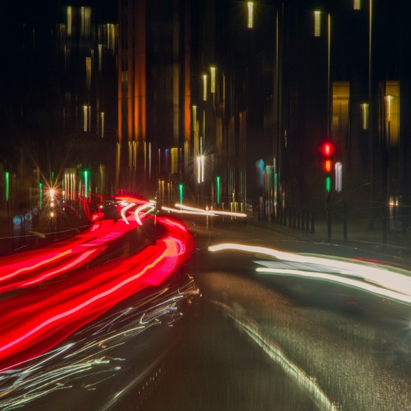 Long-exposure view down a Glasgow city street at night, with red tail-light trails curving across the left side and broad white headlight trails on the right. Red and green traffic signals glow beside dark tall buildings, and the road surface reflects the lights.