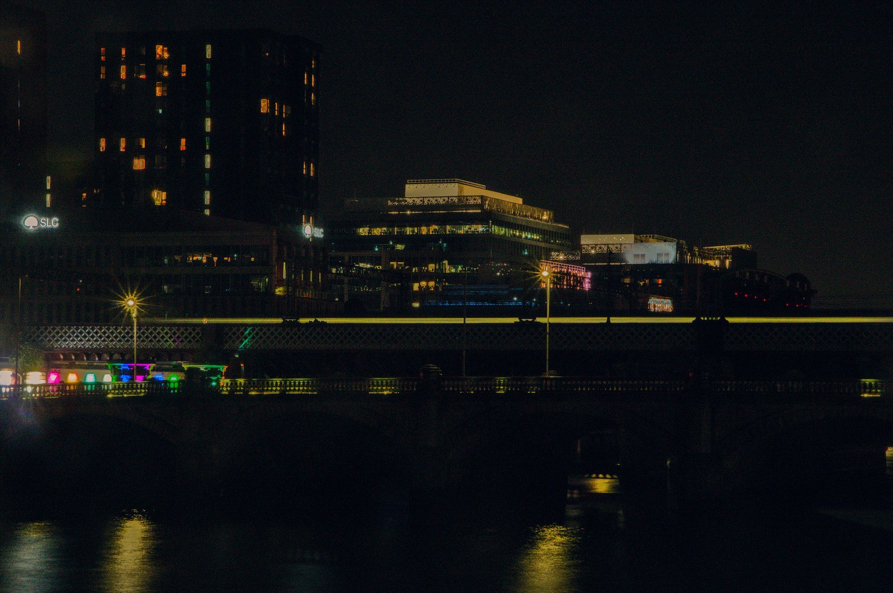 Night view across the River Clyde in Glasgow: a yellow-striped rail vehicle on a bridge or elevated track, with dark water below and office buildings with lit windows behind.