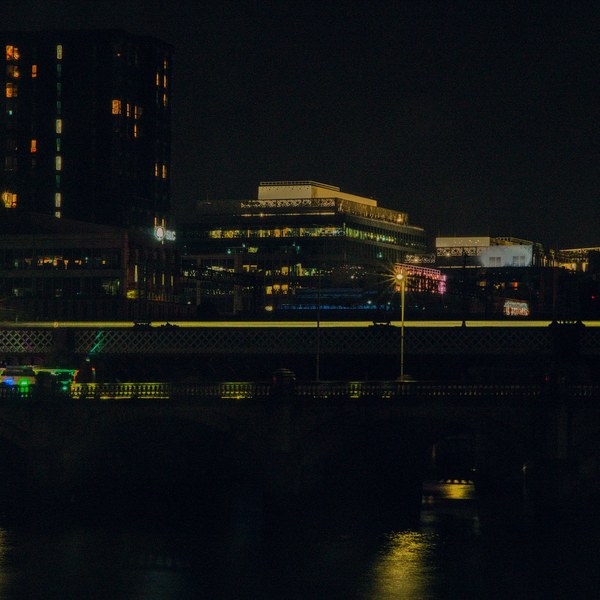 Night view across the River Clyde in Glasgow: a yellow-striped rail vehicle on a bridge or elevated track, with dark water below and office buildings with lit windows behind.