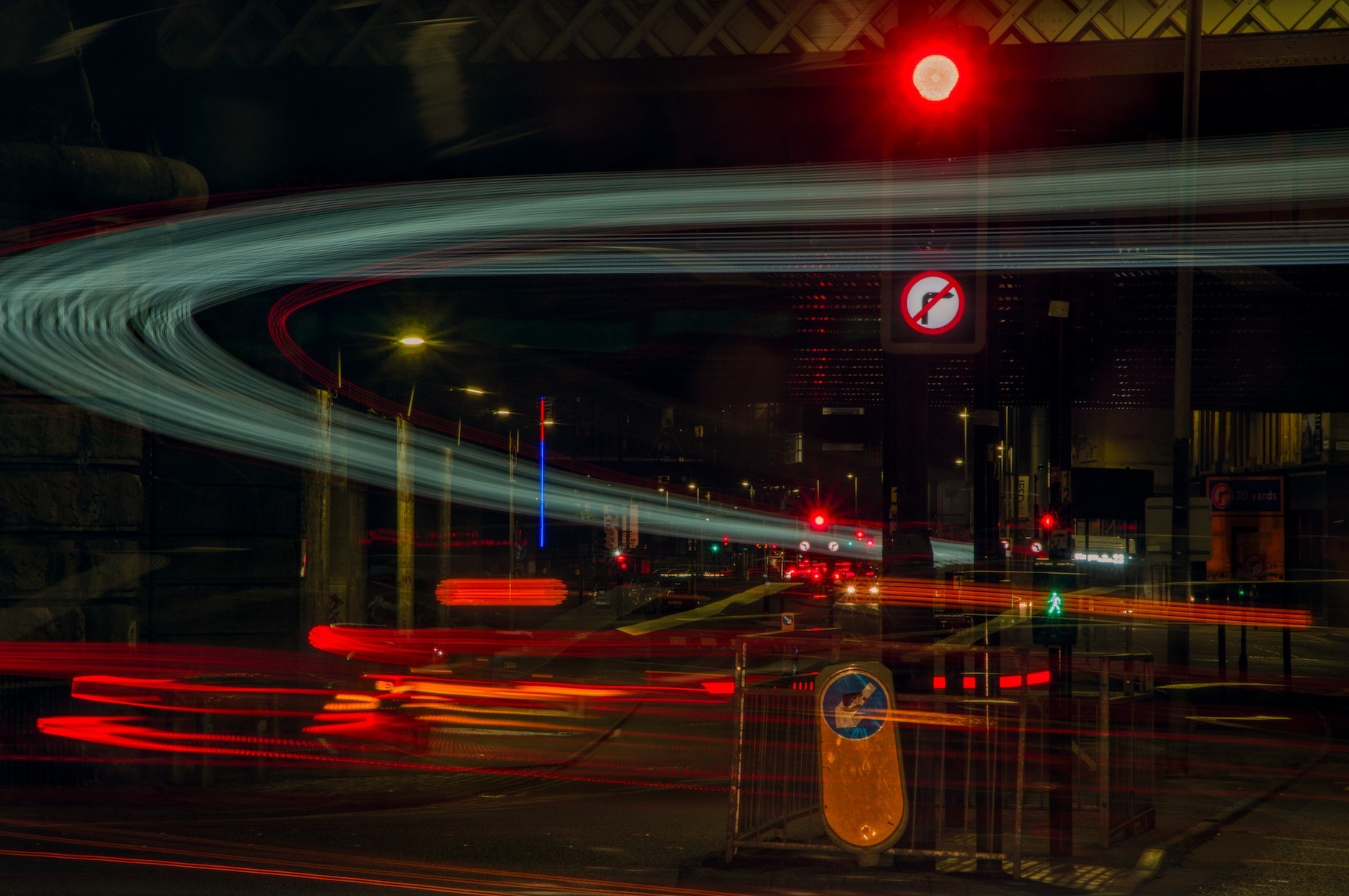 Long-exposure night view of a Glasgow road junction beneath an overhead structure, with red, amber and blue-green vehicle-light trails, a red traffic signal, road signs and pedestrian barriers.