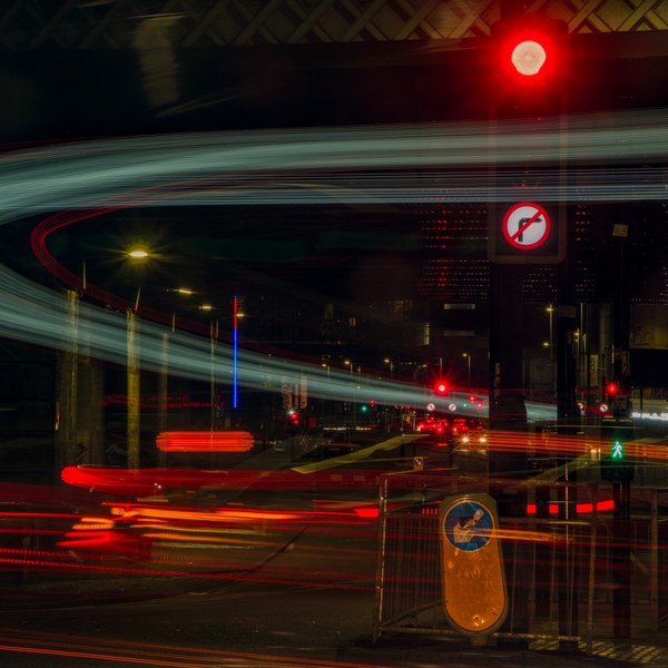 Long-exposure night view of a Glasgow road junction beneath an overhead structure, with red, amber and blue-green vehicle-light trails, a red traffic signal, road signs and pedestrian barriers.