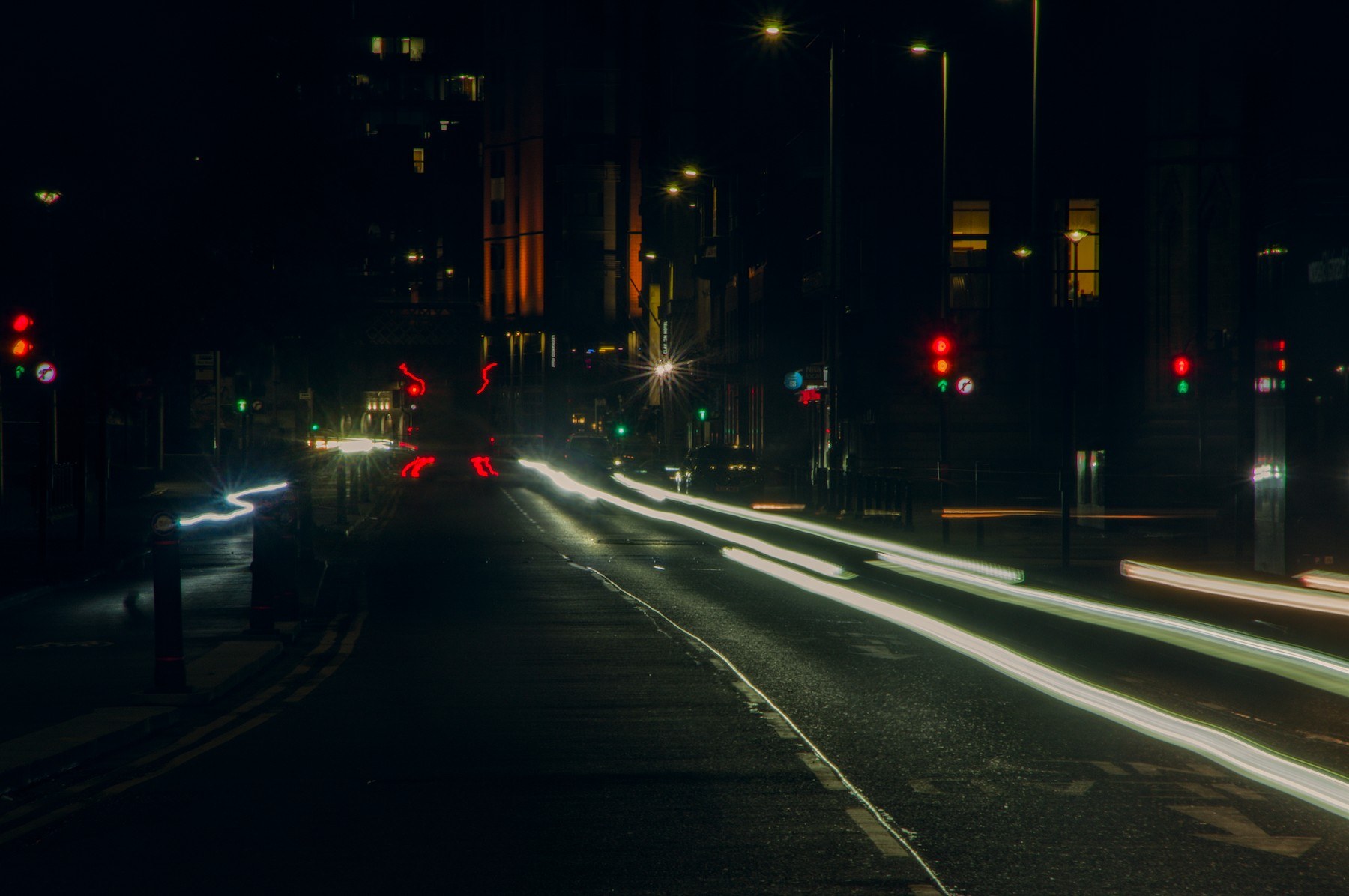 Long-exposure nighttime view down a Glasgow street, with bright white and red vehicle-light trails curving across a multi-lane road toward a signal-controlled junction. Red and green traffic lights, streetlamps, dark buildings, and faint road reflections are visible.