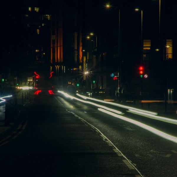 Long-exposure nighttime view down a Glasgow street, with bright white and red vehicle-light trails curving across a multi-lane road toward a signal-controlled junction. Red and green traffic lights, streetlamps, dark buildings, and faint road reflections are visible.