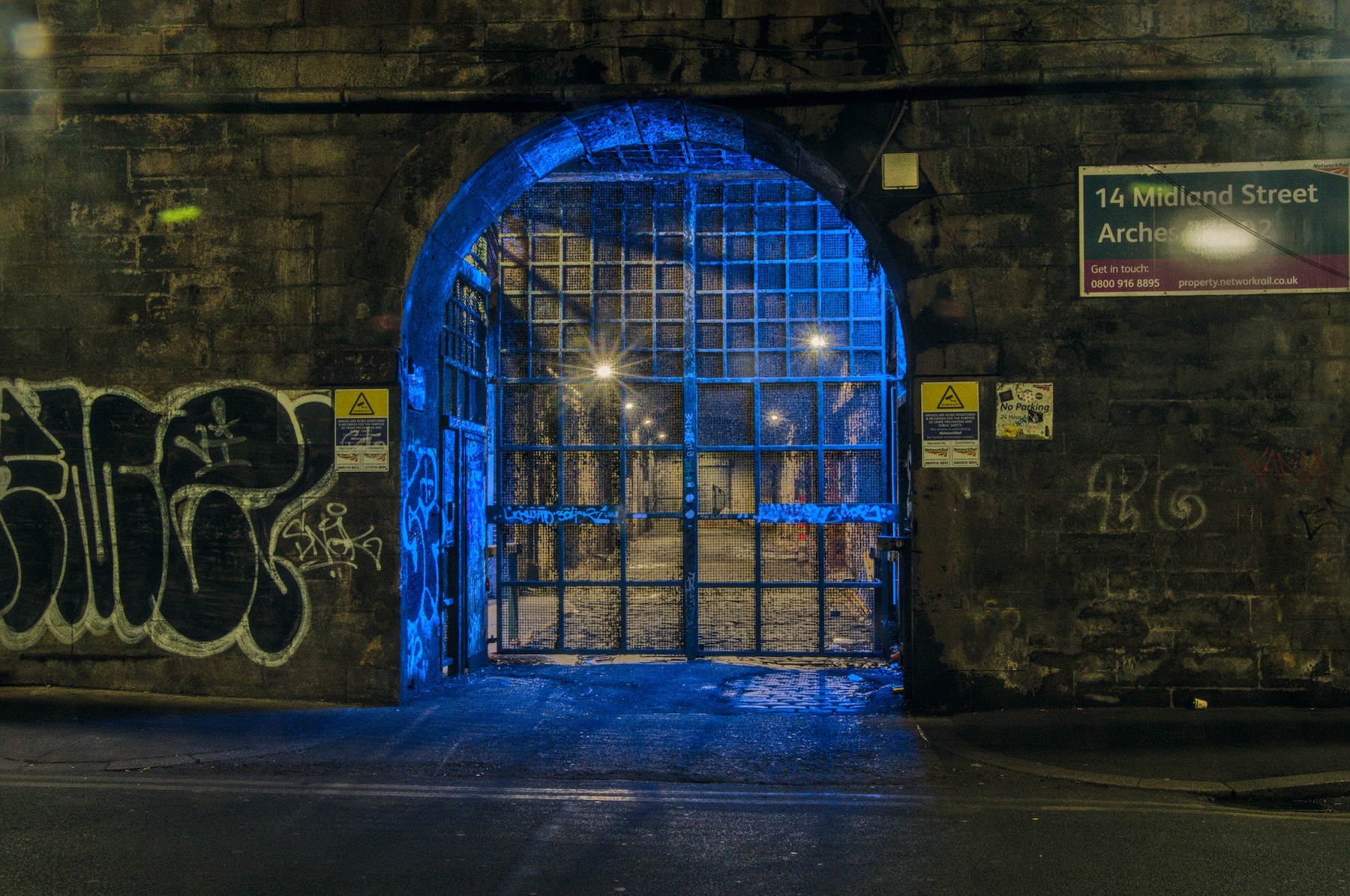Closed blue metal grid gate in a brick archway at night, with a chain across it, graffiti on both sides, warning signs near the entrance, and small warm lights visible beyond.