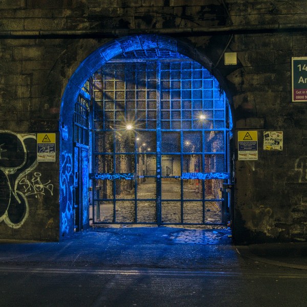 Closed blue metal grid gate in a brick archway at night, with a chain across it, graffiti on both sides, warning signs near the entrance, and small warm lights visible beyond.