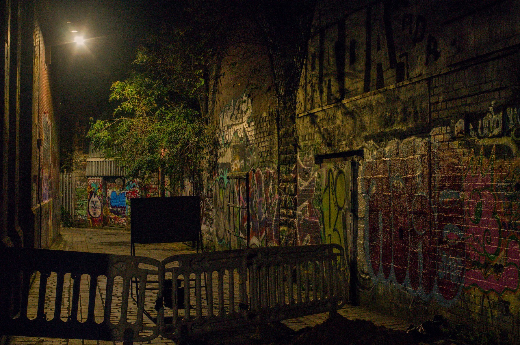 Nighttime graffiti-covered alley in Glasgow city, blocked by linked pedestrian barriers, with a black signboard, leafy foliage, and a bright streetlamp illuminating brick walls.