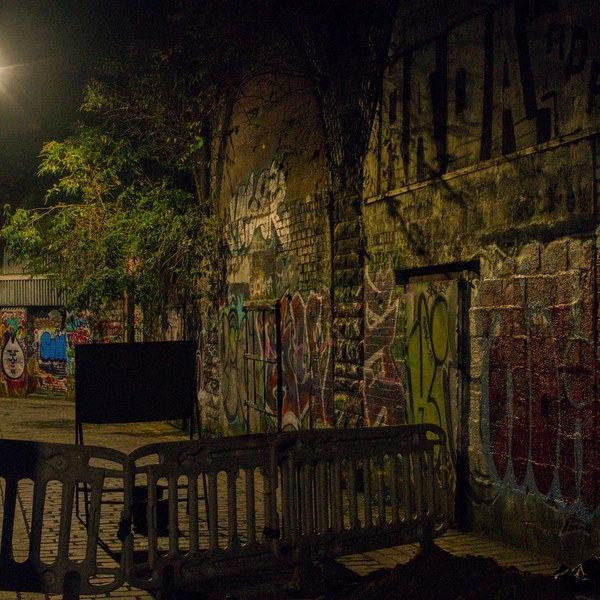 Nighttime graffiti-covered alley in Glasgow city, blocked by linked pedestrian barriers, with a black signboard, leafy foliage, and a bright streetlamp illuminating brick walls.