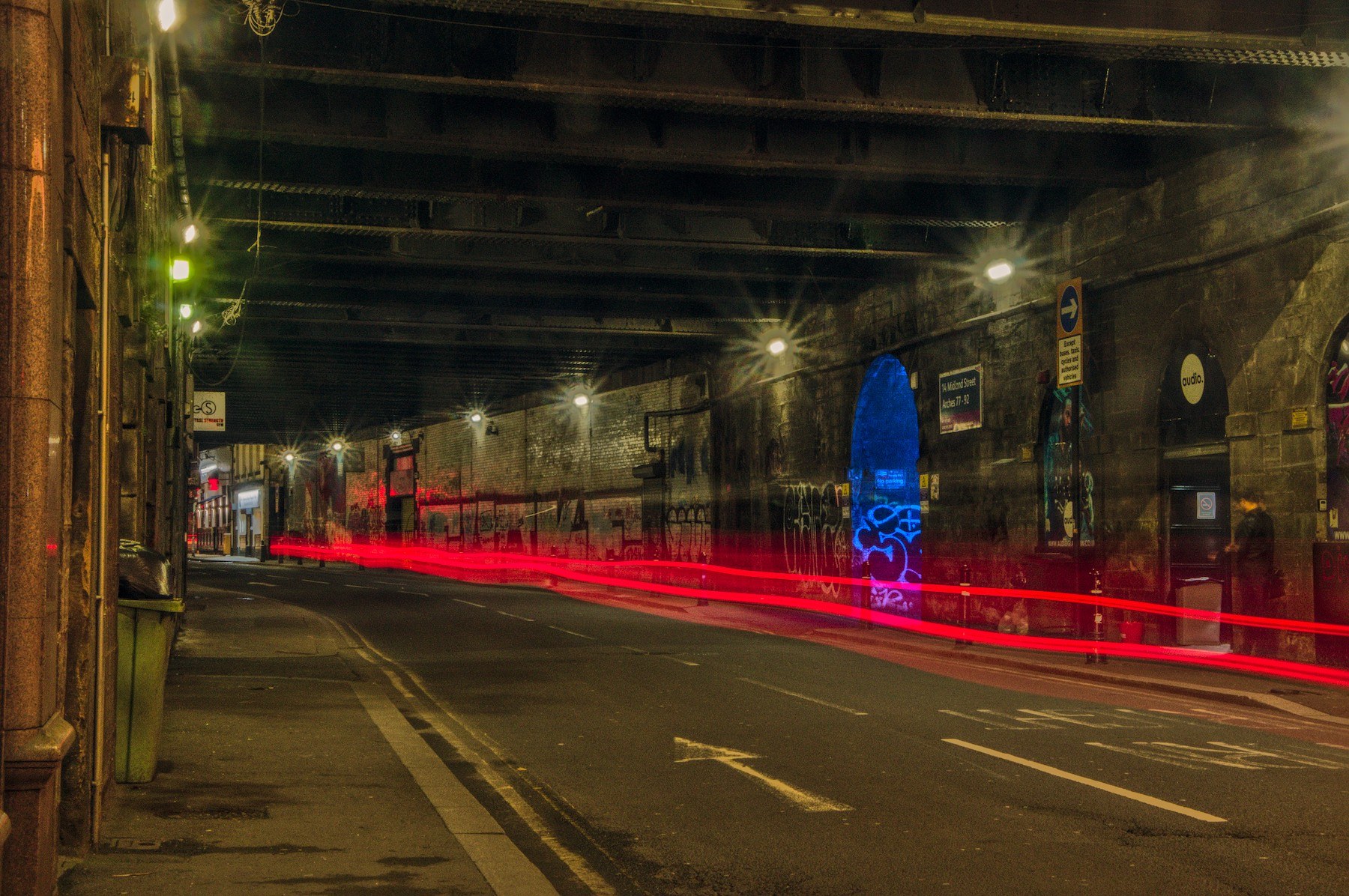 Nighttime road beneath a Glasgow viaduct, with red vehicle light trails crossing the lanes, starburst streetlights, graffiti-covered retaining walls and a bright blue arched recess on the right.