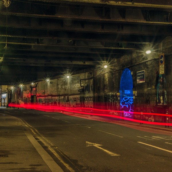 Nighttime road beneath a Glasgow viaduct, with red vehicle light trails crossing the lanes, starburst streetlights, graffiti-covered retaining walls and a bright blue arched recess on the right.