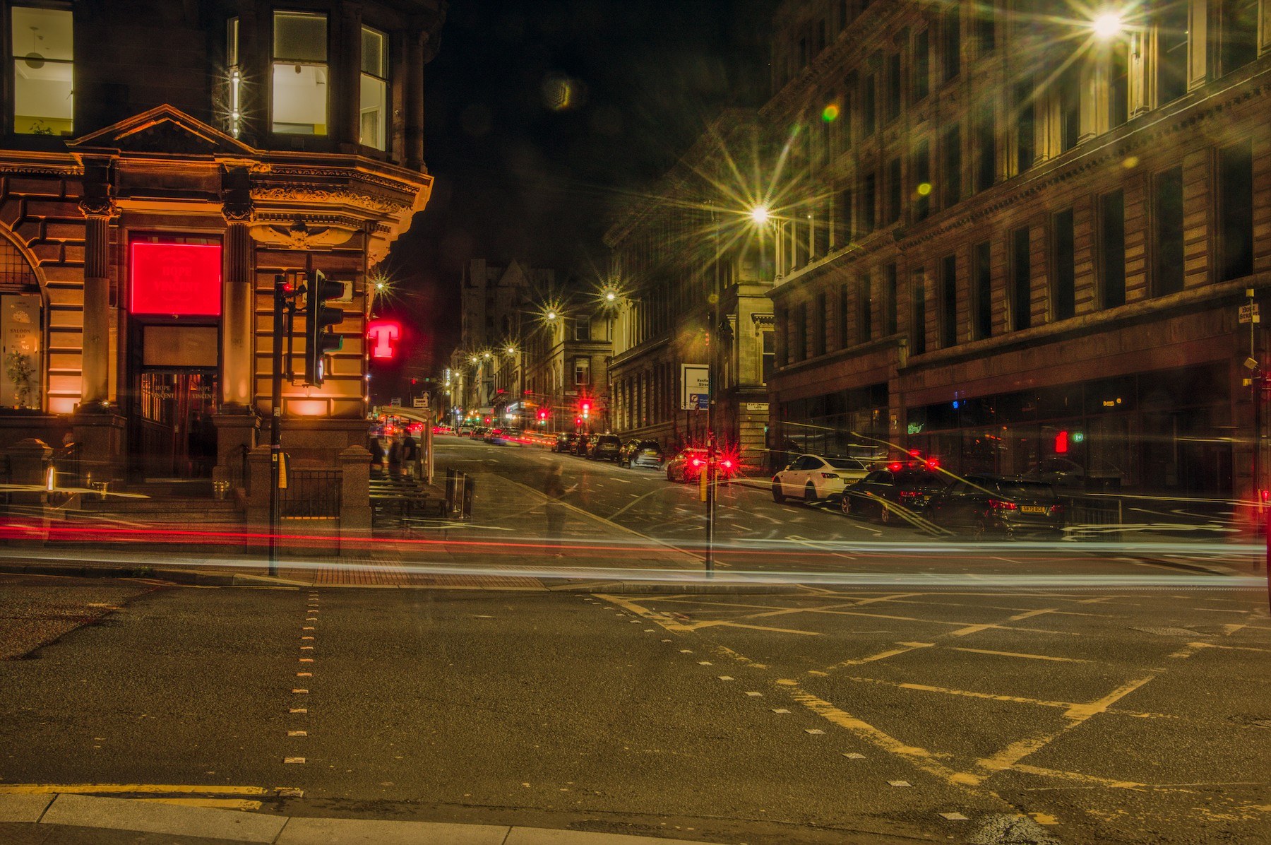 Nighttime Glasgow city-centre intersection with red and white vehicle light trails crossing the road, a warmly lit ornate corner building on the left, and a long dark building with tall windows on the right.
