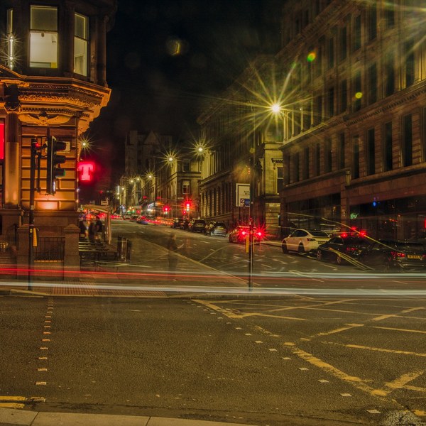 Nighttime Glasgow city-centre intersection with red and white vehicle light trails crossing the road, a warmly lit ornate corner building on the left, and a long dark building with tall windows on the right.
