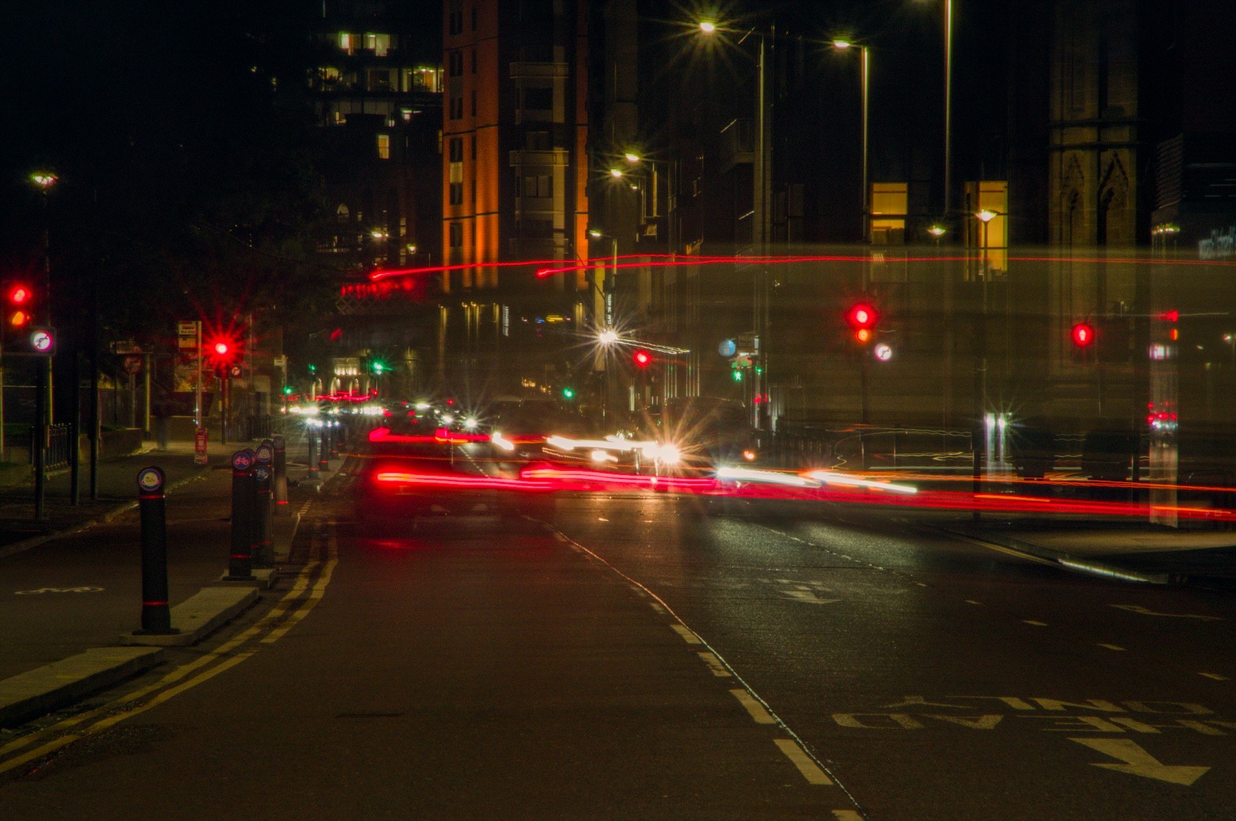 Long-exposure night view of a Glasgow road junction, with red traffic lights, red and white vehicle light trails, a blurred large vehicle crossing the road, bollards on the left pavement, and tall dark buildings.