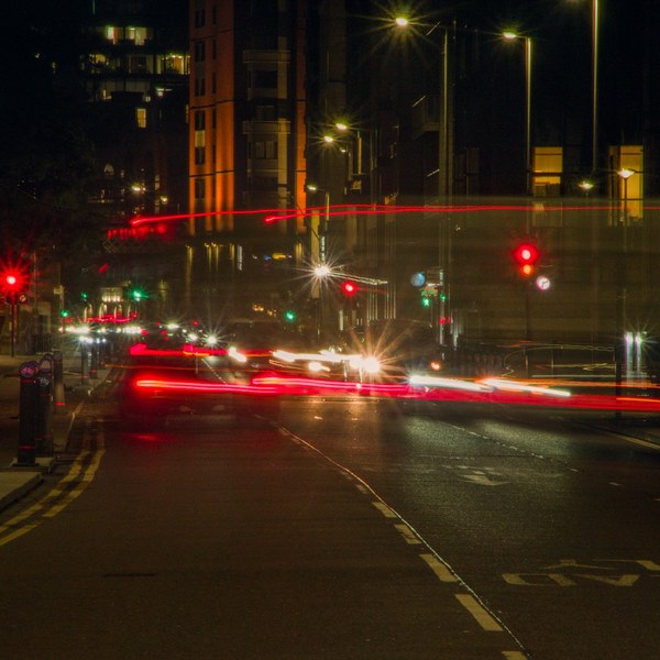 Long-exposure night view of a Glasgow road junction, with red traffic lights, red and white vehicle light trails, a blurred large vehicle crossing the road, bollards on the left pavement, and tall dark buildings.