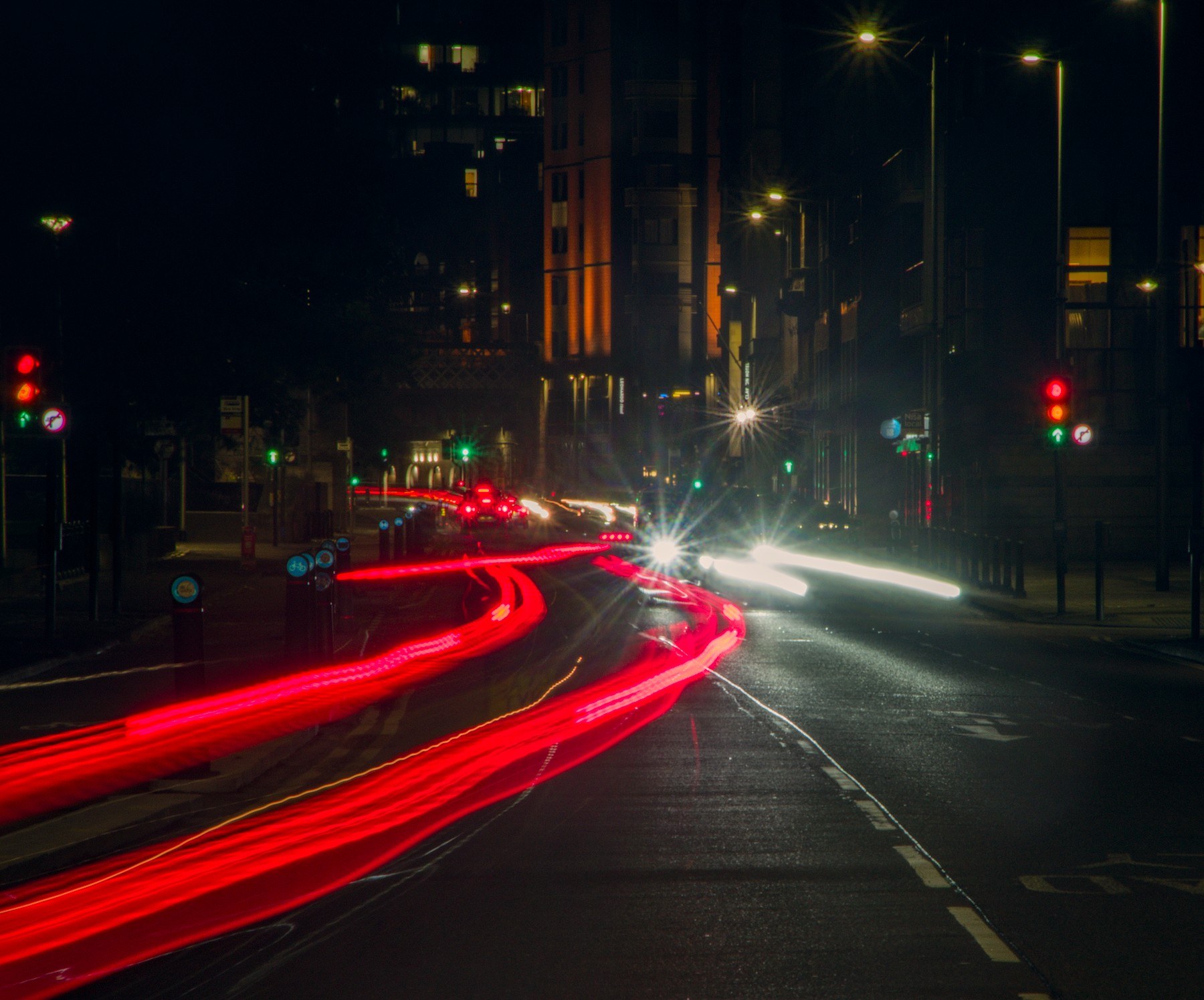 Long-exposure view of a Glasgow city street junction at night, with red tail-light trails curving from the foreground toward the centre, white headlight trails along the right, and traffic signals and streetlights between dark tall buildings.