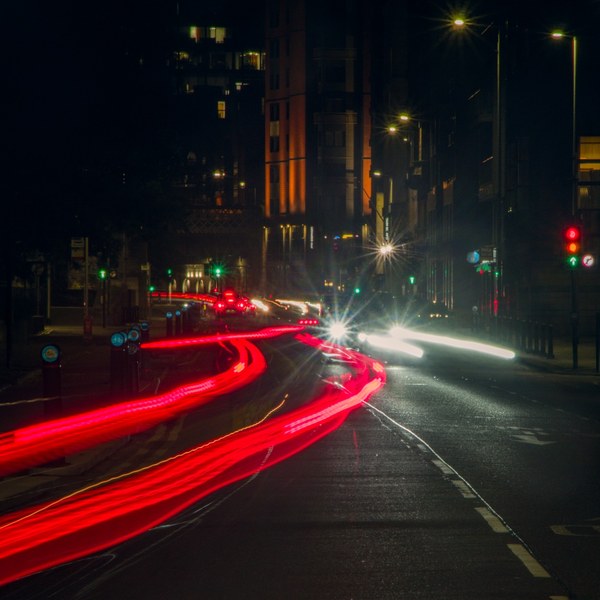 Long-exposure view of a Glasgow city street junction at night, with red tail-light trails curving from the foreground toward the centre, white headlight trails along the right, and traffic signals and streetlights between dark tall buildings.