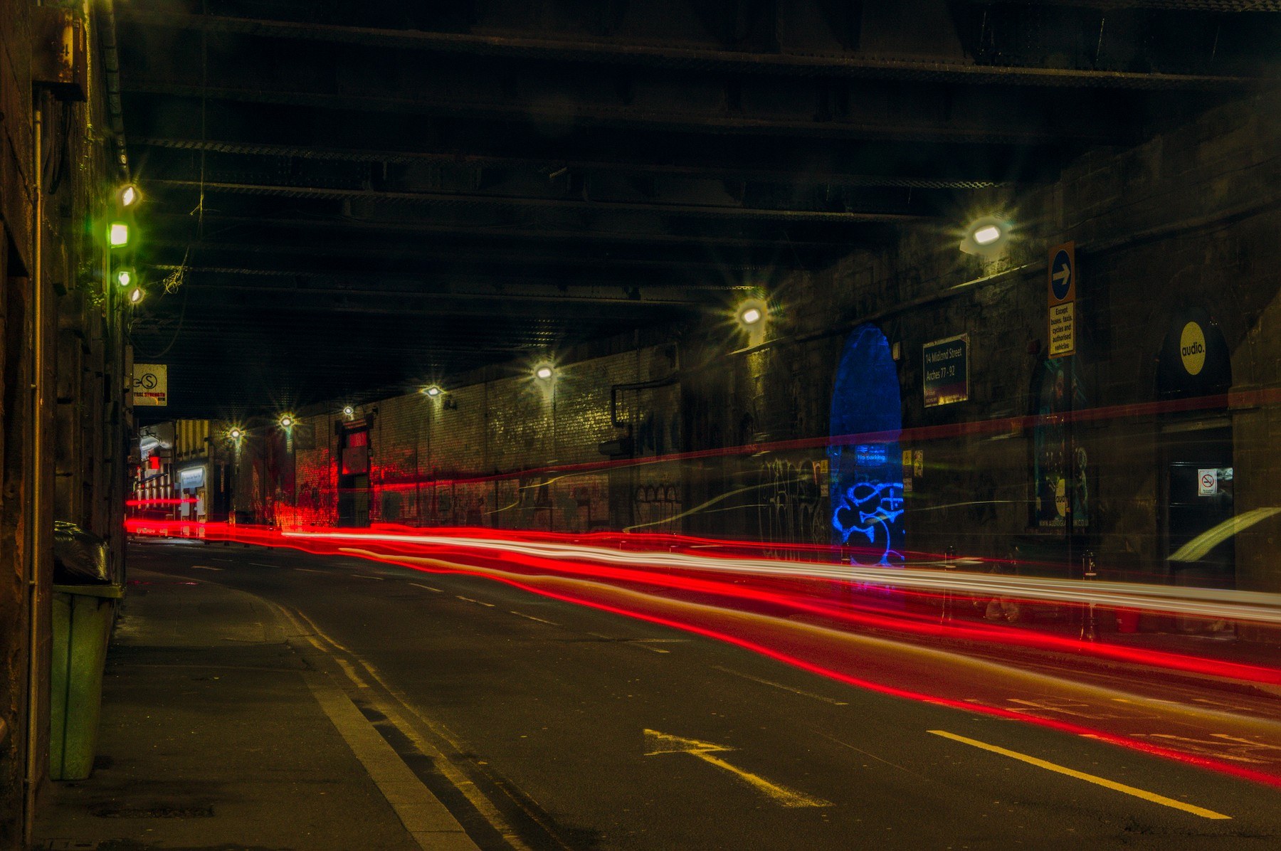 Long-exposure view along a curving Glasgow road beneath a dark brick viaduct, with red, white and orange vehicle light trails, street lamps, a narrow left pavement, and a blue graffiti-marked recess on the right wall.