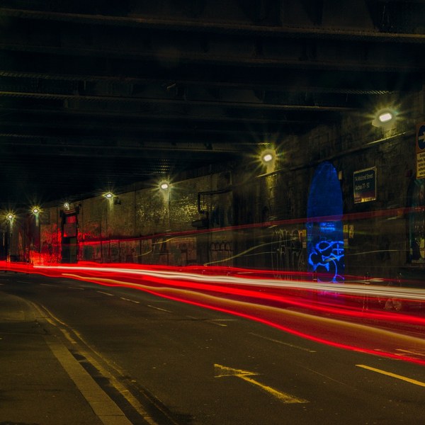 Long-exposure view along a curving Glasgow road beneath a dark brick viaduct, with red, white and orange vehicle light trails, street lamps, a narrow left pavement, and a blue graffiti-marked recess on the right wall.