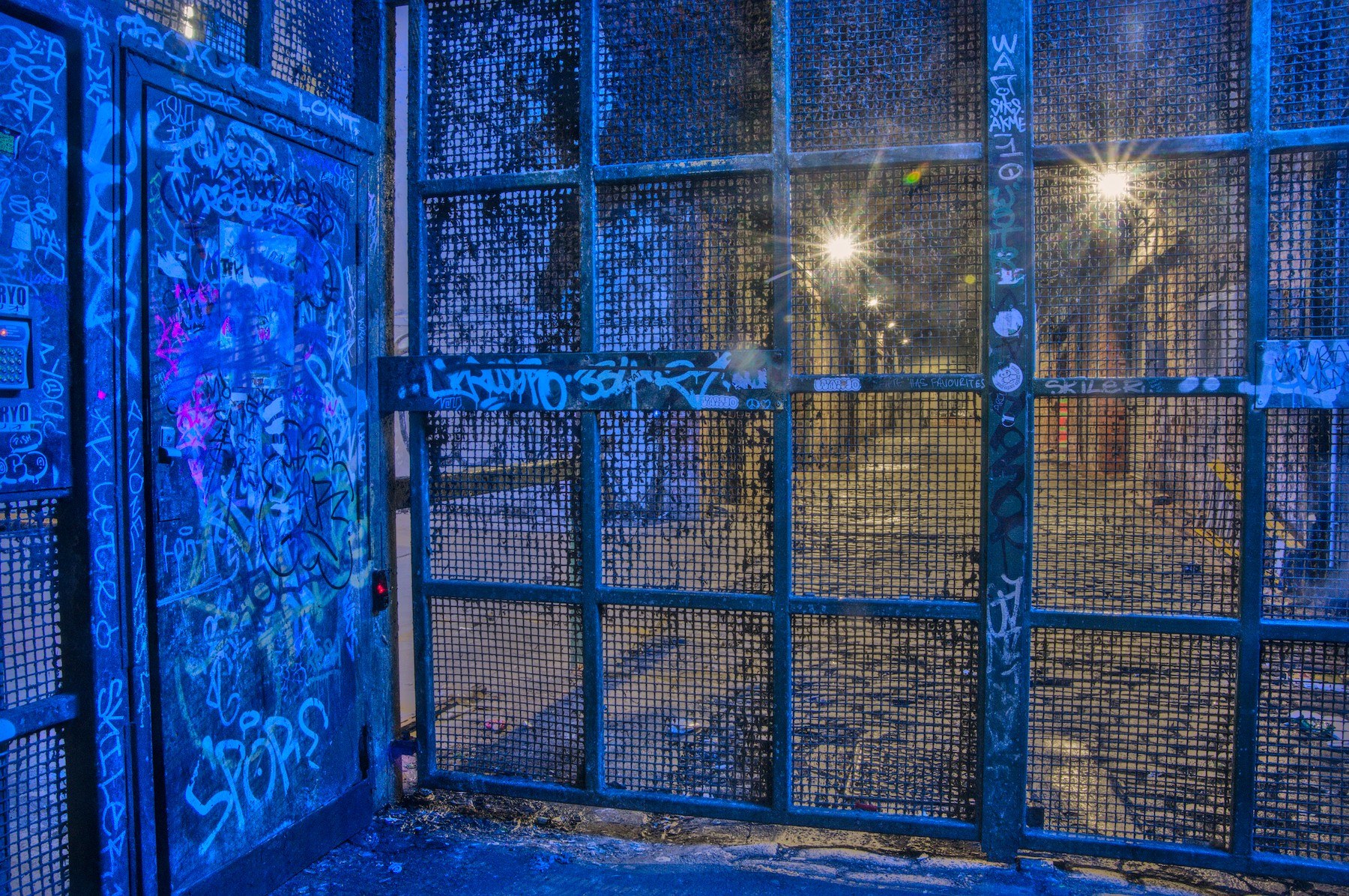 Blue-lit graffiti-covered metal door and wire-mesh security gate, with two bright lights visible through the grid behind it.