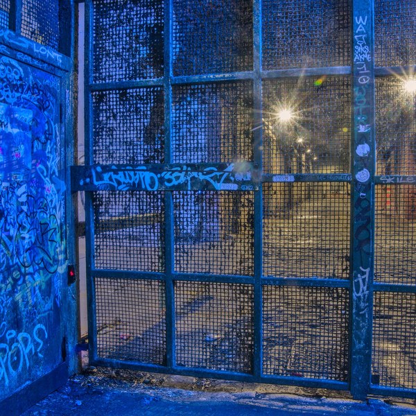 Blue-lit graffiti-covered metal door and wire-mesh security gate, with two bright lights visible through the grid behind it.