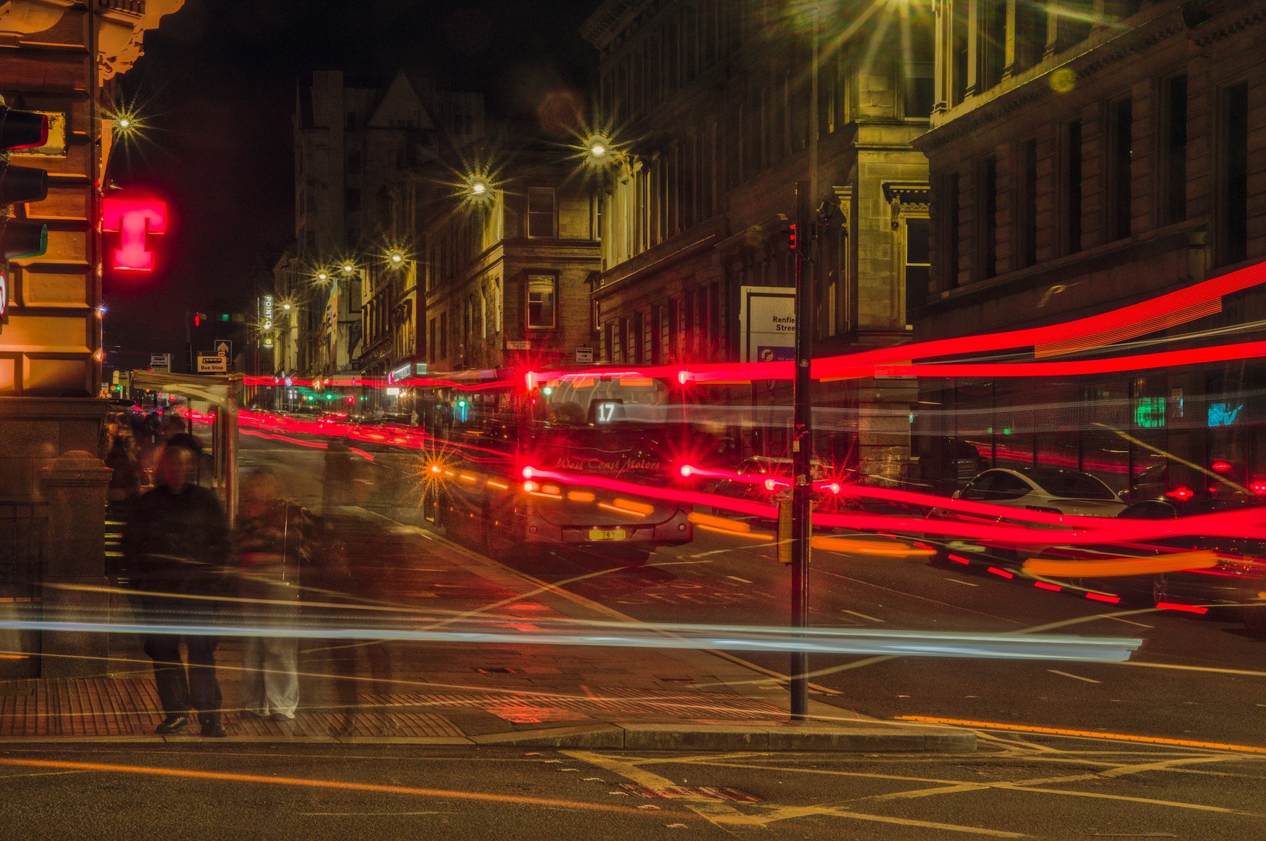Long-exposure night view of a Glasgow intersection: a bus displaying route 17 stands near the centre as red, white and blue vehicle-light trails cross the road; blurred pedestrians wait on the left beside tall stone buildings.