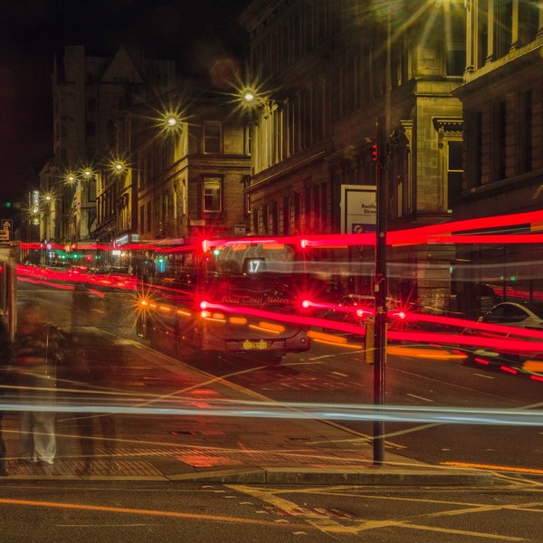 Long-exposure night view of a Glasgow intersection: a bus displaying route 17 stands near the centre as red, white and blue vehicle-light trails cross the road; blurred pedestrians wait on the left beside tall stone buildings.